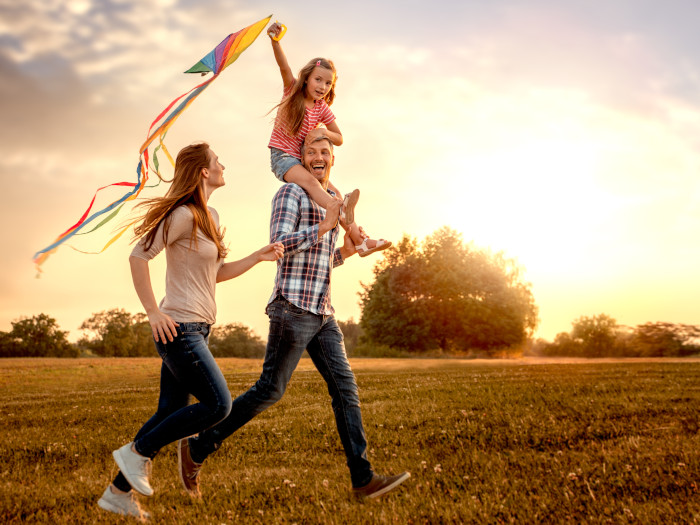 Una familia de tres corre sobre la hierba, una niña se sienta sobre sus hombros con una cometa en las manos