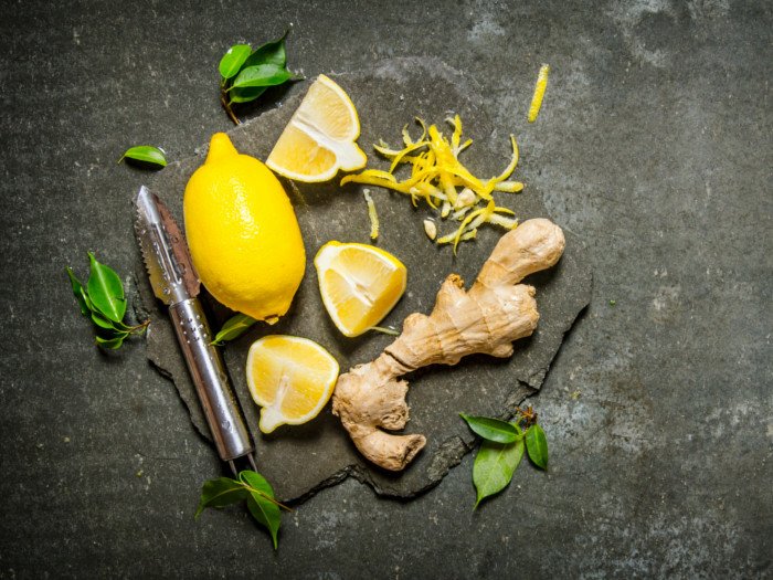 Vista plana de limón entero, limón en rodajas, jengibre, pelador y hojas de limón en la superficie de pizarra.