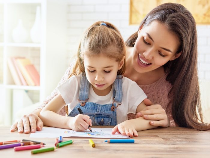 Una mujer sonríe mientras se sienta detrás de una chica que está coloreando su libro para colorear