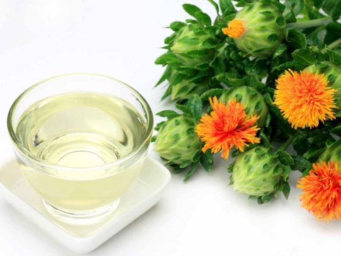 Un vaso de aceite de cártamo junto a flores rojas sobre un fondo blanco.